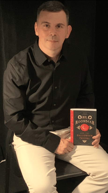 A person sitting on a chair, holding a book titled 'Mr. Moondream and the Halloween Crystal', wearing a black shirt and white pants.